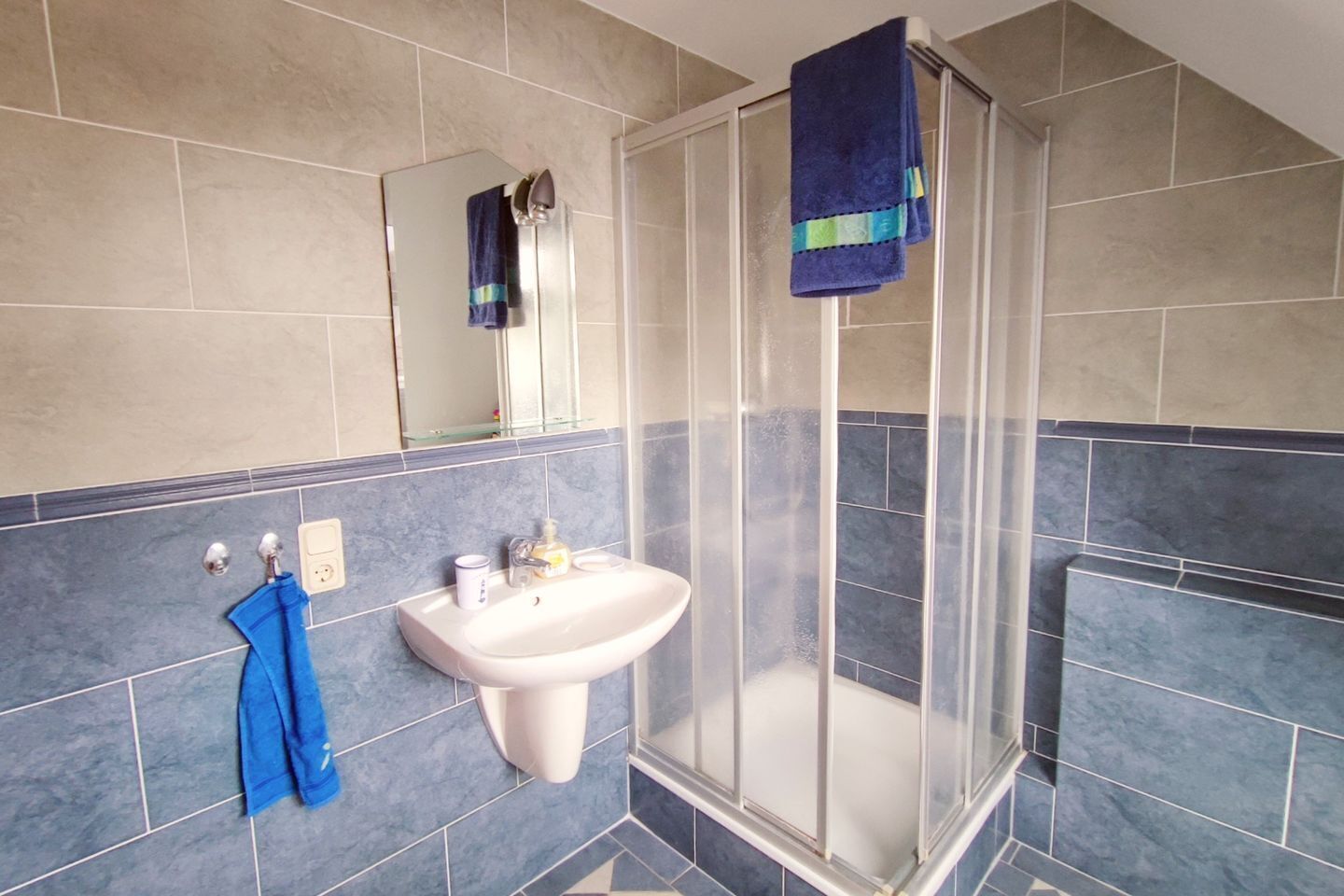 Un baño con lavamanos blanco, cabina de ducha con mampara de vidrio y azulejos en tonos azul y beige.