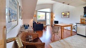 A modern living room with a sofa, dining table, and chairs, a kitchen area with a dishwasher, and a large window providing natural light.