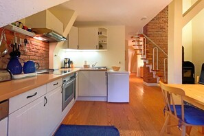 A compact kitchen with white cabinets, a stove, and a refrigerator. There is a wooden dining table with a blue chair. A brick wall and a staircase with a metal railing are visible in the background. - Hochwertig, Moderne und Gemütliche Ferienwohnung mit 55qm für Vier Personen. - Blau-weiss (Loddin)