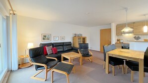 A modern living room with a sofa, chairs, a coffee table, and a dining table. There is a kitchen area with cabinets and a stove.