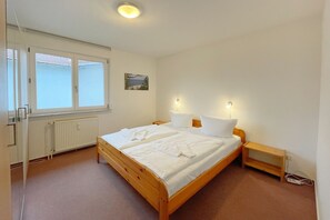 A bedroom with a wooden bed, white bedding, a radiator, a window with blinds, and a small bedside table with a lamp. - "Relaxation and comfort: Your vacation apartment on Usedom awaits you!" - (Loddin)
