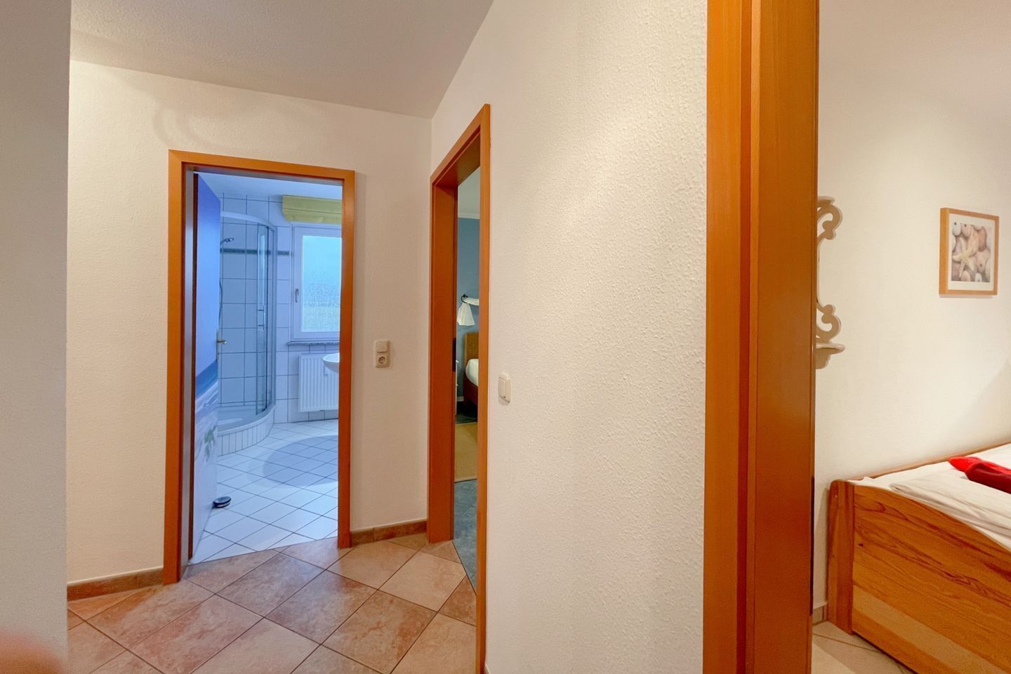 A hallway leading to a bathroom with a shower and a toilet. There is a bed with a red blanket in the room.