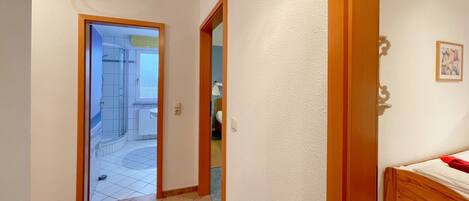 A hallway leading to a bathroom with a shower and a toilet. There is a bed with a red blanket in the room.
