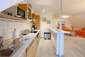 Una cocina moderna con armarios de madera, un microondas, un tostador y una cafetera. Hay un cubo de basura y una mesita con una silla. La cocina da paso a una zona de estar con un sofá y un cuadro en la pared.