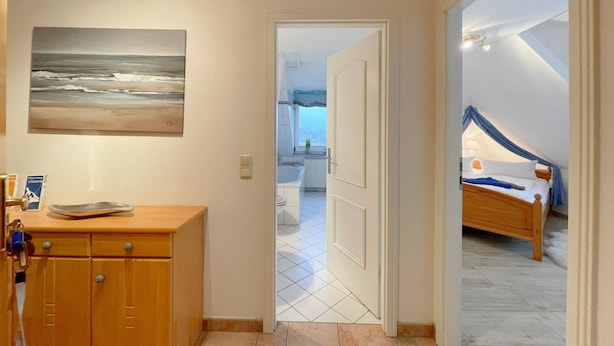 A hotel room with a wooden dresser, a painting of a beach, and a view of the sea through an open door.