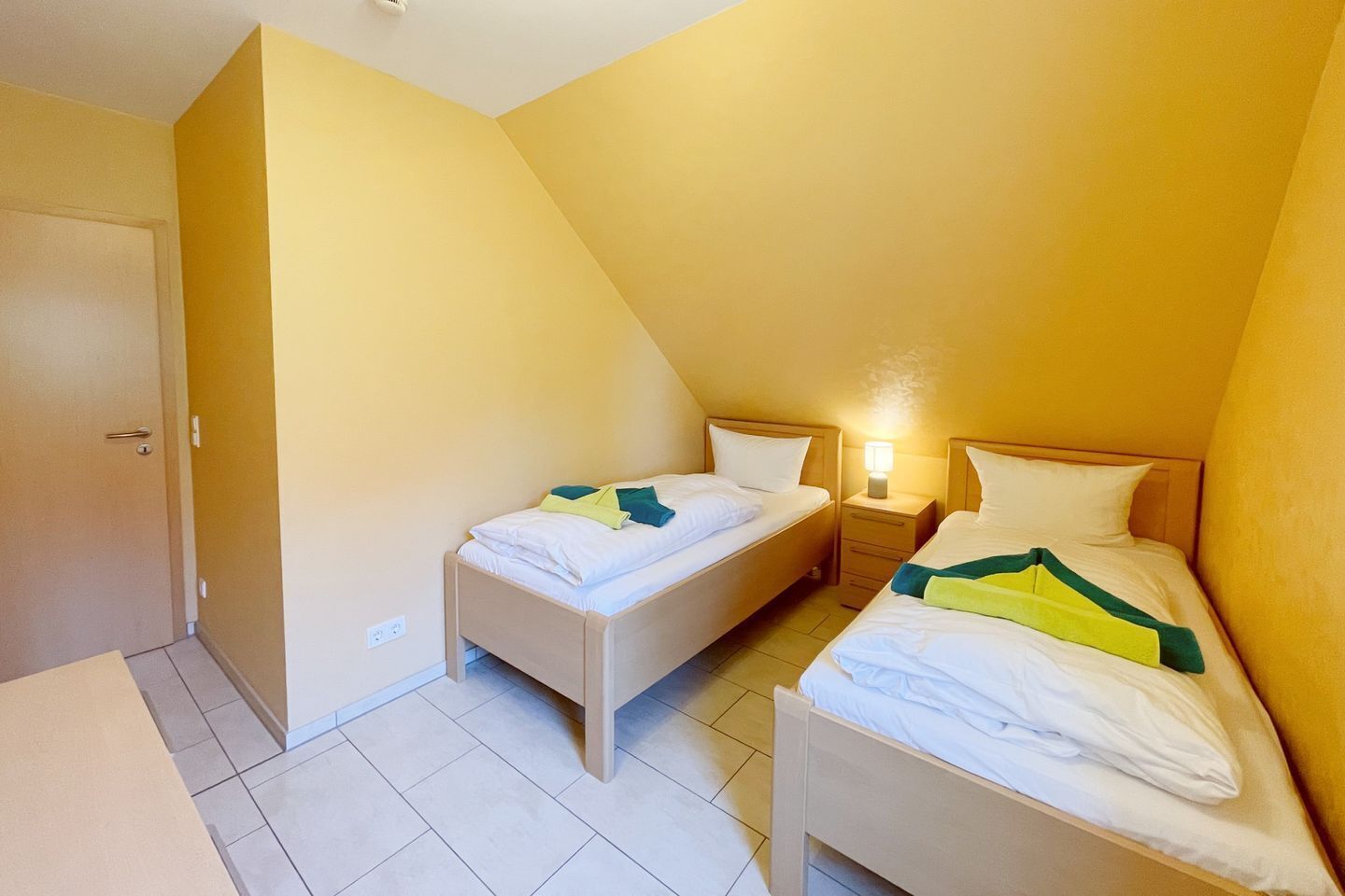 A small, triangular room with two single beds, a bedside table with a lamp, and a door.