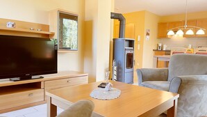 A cozy living room with a flat-screen TV, a dining table, and comfortable seating. A modern kitchen with a stove and cabinets is visible in the background.