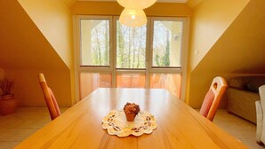 A dining room with a wooden table, a cupcake on a doily, and a view of trees through large windows.