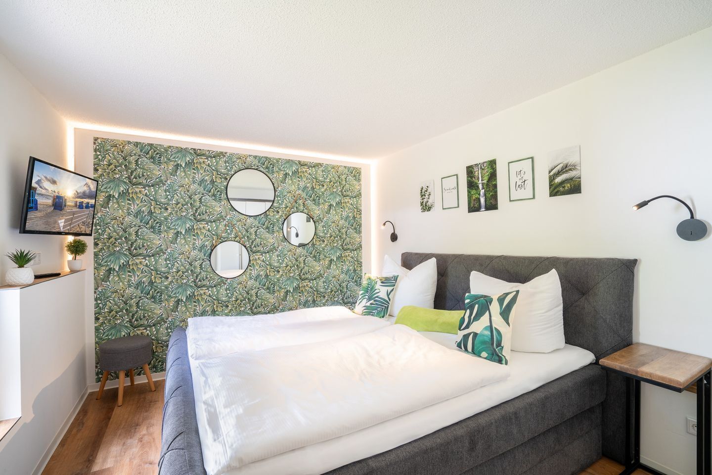 A modern bedroom with a large bed, a grey upholstered headboard, and a wall adorned with mirrors and framed pictures.