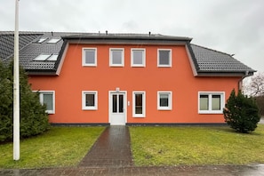 A two-story building with a red facade, a central entrance with a door marked '7C', and multiple windows. - Liebevoll Eingerichtete, 55 qm Große, Ferienwohnung für 3 Personen Geeignet. - Cölpinstrasse 7c (Loddin)