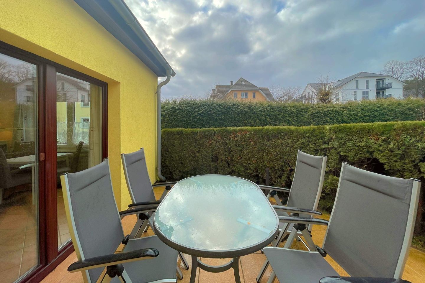 Eine Terrasse im Freien mit einem Glastisch und vier Stühlen, einer gelben Wand und einer Hecke im Hintergrund.