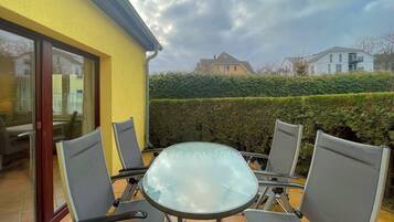 Eine Terrasse im Freien mit einem Glastisch und vier Stühlen, einer gelben Wand und einer Hecke im Hintergrund.