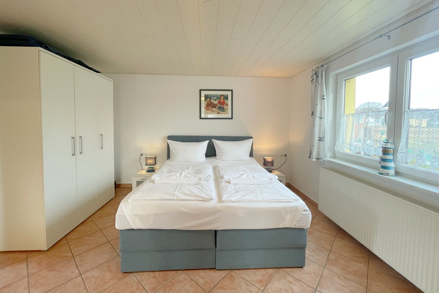 A bedroom with a bed, two bedside tables with lamps, a wardrobe, a window with curtains, and a small decorative item on the windowsill.