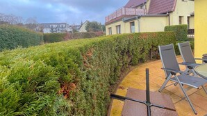 En terrasse med stole og et bord, omgivet af pænt beskåret hække og en gul bygning med rødt tag.