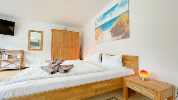 A bedroom with a wooden bed, white bedding, a bedside table with a lamp, a TV on the wall, and a painting of a beach scene. - "Experience unforgettable days in the "Engel" vacation bungalow on the Baltic Sea!" - To (Ückeritz)
