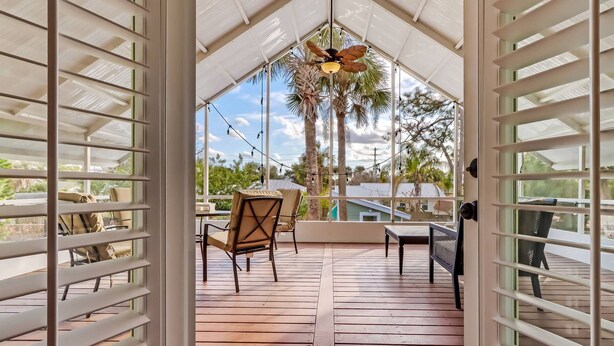 A covered outdoor patio with wooden flooring, a ceiling fan, and a view of palm trees and a house. - Florida Breeze (St. Augustine)