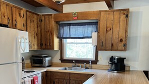 Une cuisine avec des meubles en bois, un réfrigérateur blanc, un four à micro-ondes, un évier et une machine à café. Il y a une fenêtre au-dessus de l’évier et un plafond en bois avec des poutres apparentes.