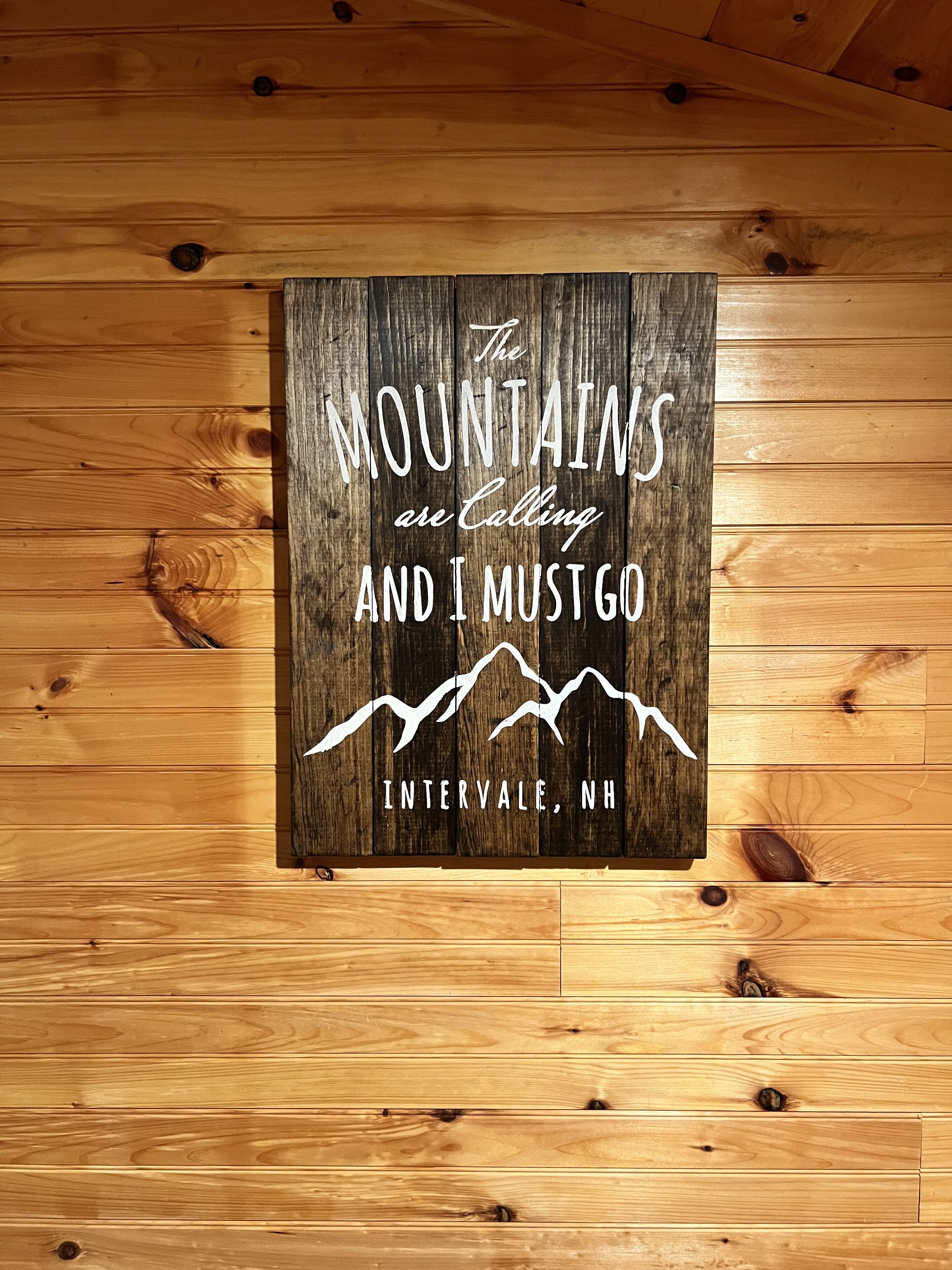 A wooden sign with a mountain graphic and text stating "The Mountains are Calling and I Must Go, Intervale, NH."