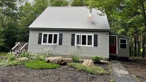 Une petite maison à un seul étage, avec un toit en métal, entourée d’un jardin où poussent différentes plantes et où se trouve un grand rocher.