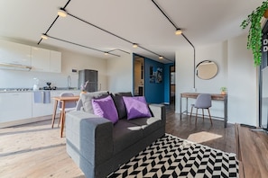 Un appartement moderne avec un canapé gris, un tapis noir et blanc, un coin repas et une cuisine dotée de meubles blancs.