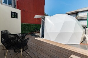 Eine Dachterrasse mit einer weißen geodätischen Kuppel, schwarzen Metallstühlen und einem roten Gebäude im Hintergrund.