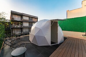 Eine kugelförmige Geodätkuppel auf einer Dachterrasse mit Holzdeck, umgeben von einem grünen Maschendrahtzaun und einem Gebäude mit Balkonen.