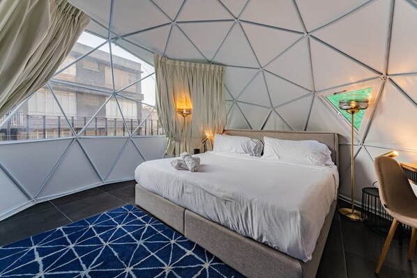 Ein modernes Hotelzimmer mit einem geometrischen Deckenmuster, einem großen Bett mit weißen Leinen, einem blau gemusterten Teppich und Blick auf die Stadt durch große Fenster.