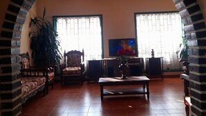 A living room with a brick archway, two windows with blinds, a television, a table with a vase, and a sofa. - Hotel Casa san rafael  (Villa de Leyva)