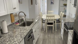 Une cuisine dotée de plans de travail en granit, d’armoires blanches, d’une table à manger avec des chaises, d’un ventilateur de plafond et d’un réfrigérateur.