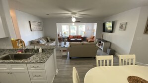 Une cuisine moderne dotée d’un comptoir en granit et d’un évier en acier inoxydable, un coin repas avec une table et des chaises blanches, et un salon avec un canapé et une table basse.