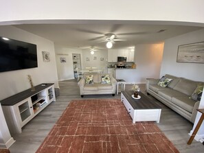 A modern living room with a flat-screen TV, a sofa, a coffee table, and a kitchen in the background.