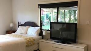 A bedroom with a bed, a TV on a stand, a window with a view of greenery, and a nightstand with a lamp. - Large one bedroom Cottage in the heart of St Marys (Saint Marys)