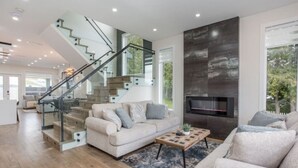 A modern living room with a staircase, a fireplace, and a large window. - Gatsby`s Corner | King Bed | Near Parks & Golf (Surrey)