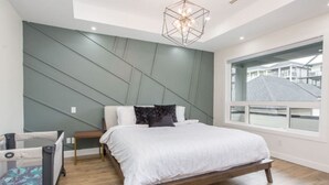 A modern bedroom with a bed, a nightstand, a chair, and a window with a view of buildings. - Gatsby`s Corner | King Bed | Near Parks & Golf (Surrey)