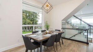 A modern dining area with a wooden table, dark chairs, and a hanging light fixture. - Gatsby`s Corner | King Bed | Near Parks & Golf (Surrey)