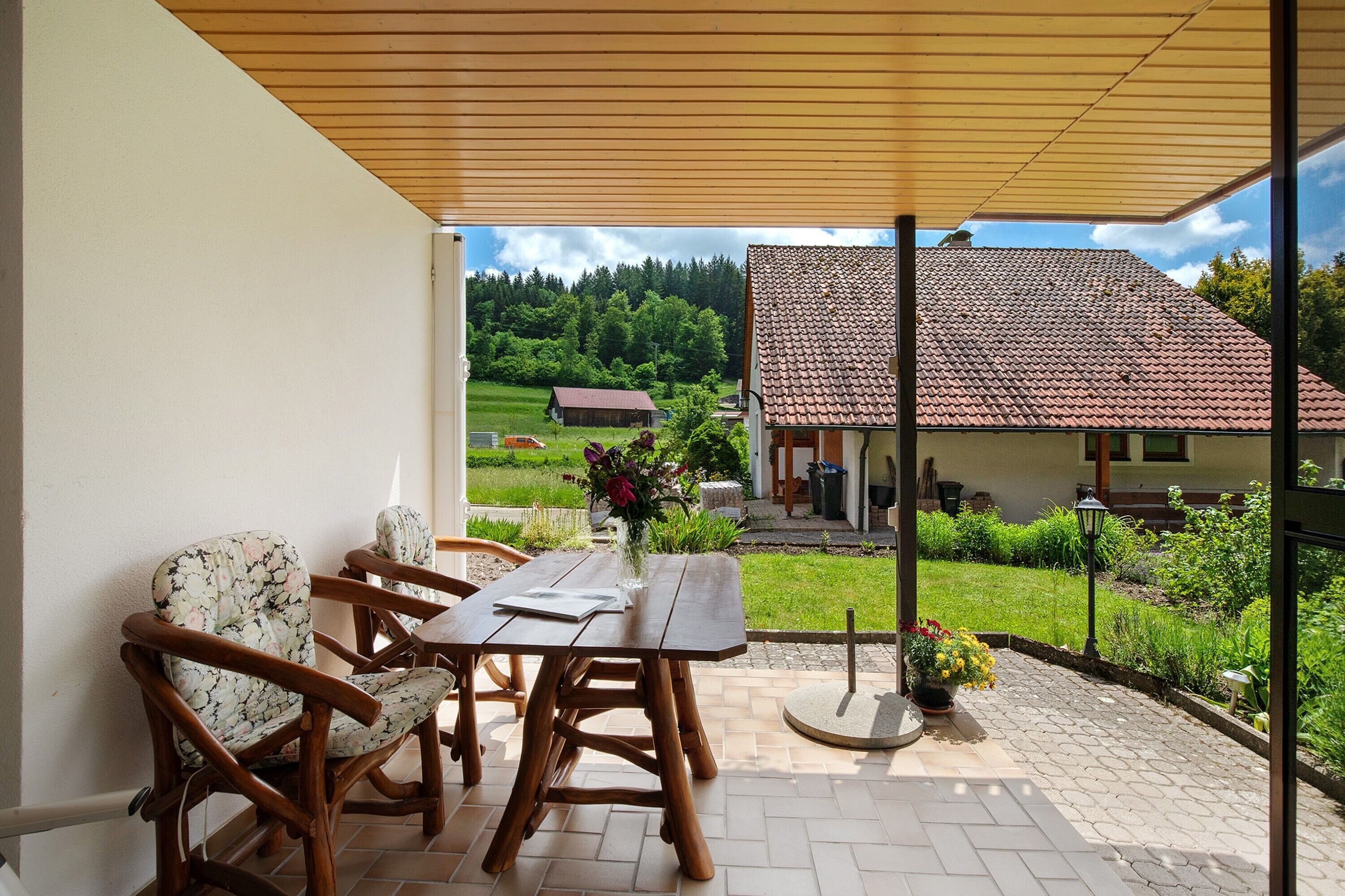 A patio with a wooden table and chairs, a potted plant, and a view of a house with a tiled roof.