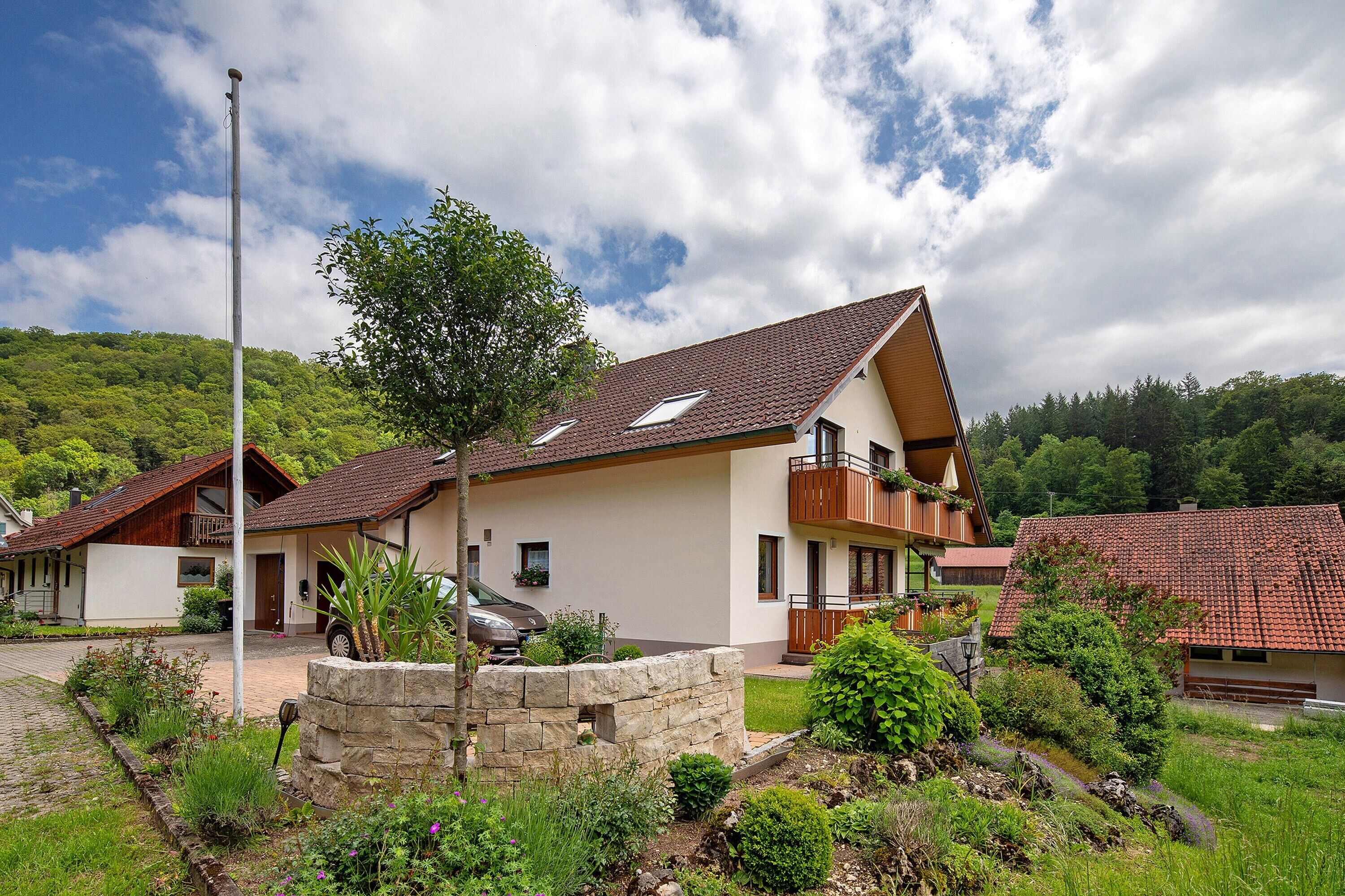 A two-story house with a sloped roof, a balcony, and a stone wall in the front garden.