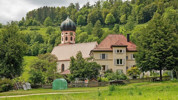 Eine Kirche mit einer markanten Kuppel, ein Wohngebäude mit rotem Dach und ein üppiger, grüner Hügel.