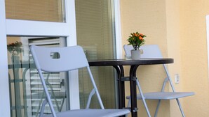 A balcony with two white chairs and a small table, a potted plant, and a window with blinds.