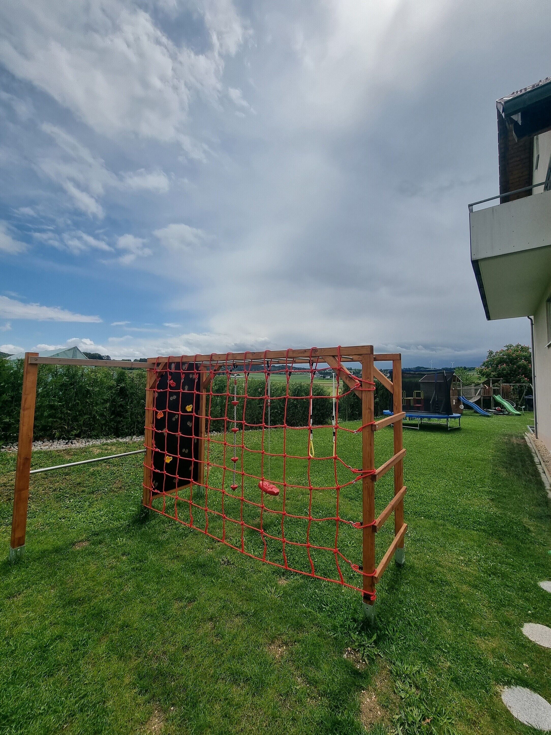 Une aire de jeux avec une structure d’escalade en forme de filet, un toboggan et un trampoline.