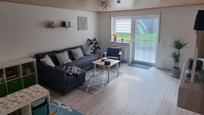 A living room with a sofa, a small table, a bookshelf, and a sliding glass door leading to a balcony. - Holiday Apartment "Biosphärenblick" with Private Terrace, Shared Garden and Wi-Fi (Münsingen)