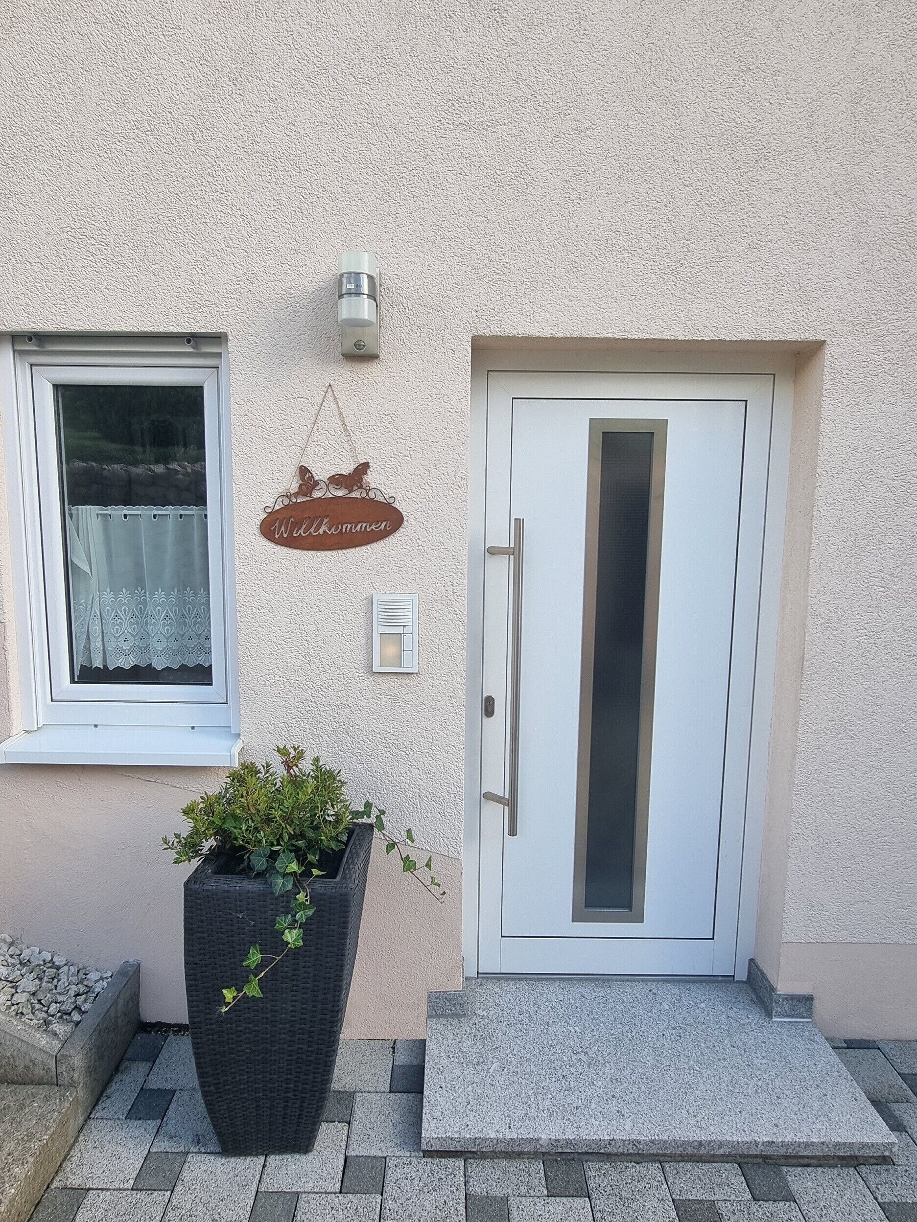 A building entrance with a white door, a window with a lace curtain, a welcome sign, and a potted plant.