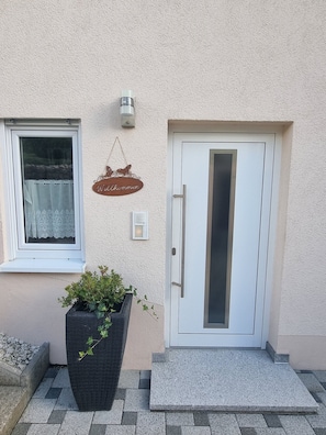 Ein Gebäudeeingang mit weißer Tür, einem Fenster mit Spitzendecke, einem Willkommensschild und einem Blumentopf.