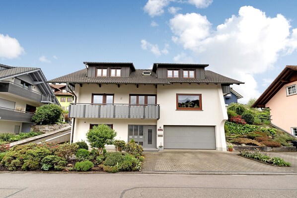 A two-story residential building with a grey roof, a balcony, and a garage.
