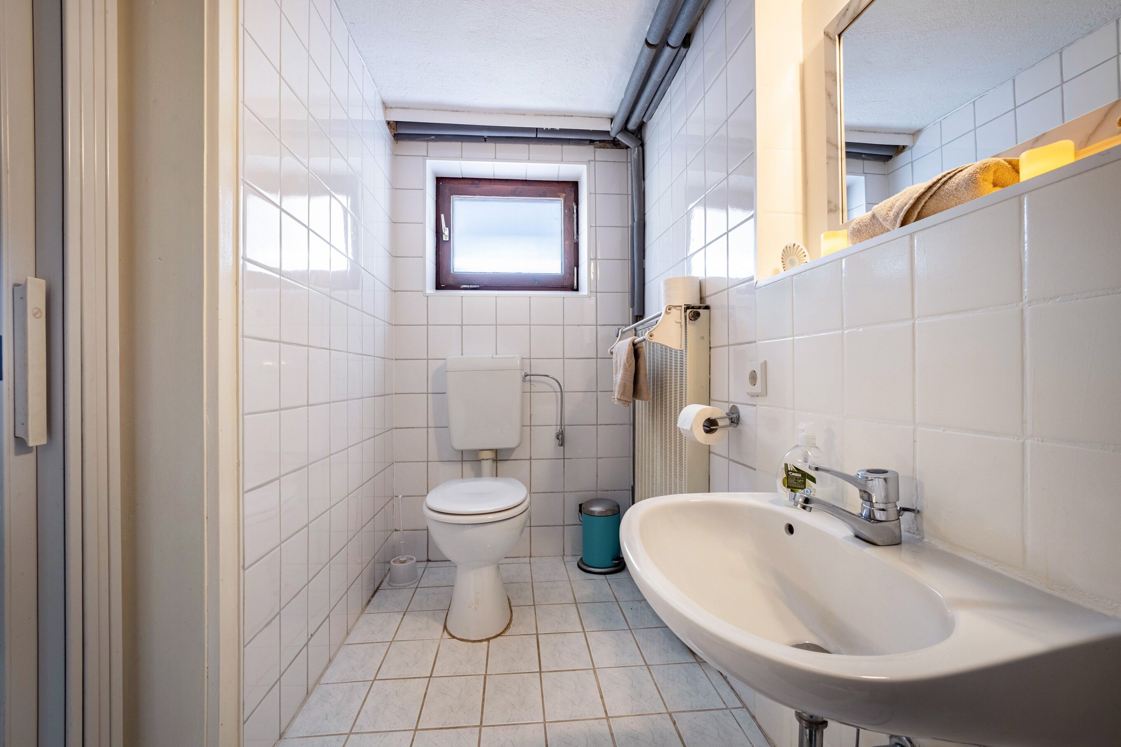 A small bathroom with white tiles, a toilet, a sink, a window, and a mirror.