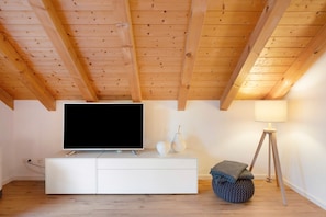 Un salon moderne avec un plafond en bois, un meuble de rangement blanc pour l’équipement audiovisuel, un téléviseur à écran plat, une lampe sur pied et un pouf rond.