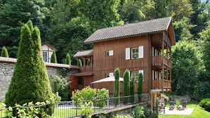 Une maison en bois dotée d’un balcon, entourée de verdure et d’un mur en pierre.