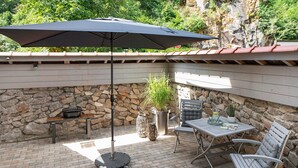 Una veranda esterna con tavolo, sedie, barbecue, una pianta in vaso e un muro in pietra.