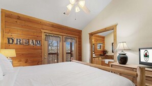 A bedroom with a wooden headboard, a bed with white bedding, a ceiling fan, a mirror, and a bedside table with a lamp.