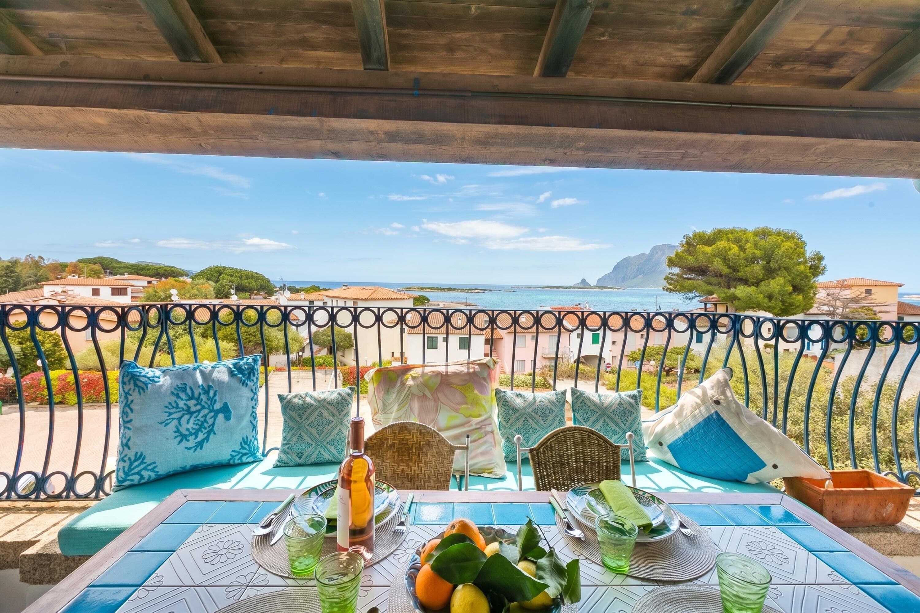A table set with a bottle, glasses, and fruits, overlooking a coastal view with a railing and chairs.